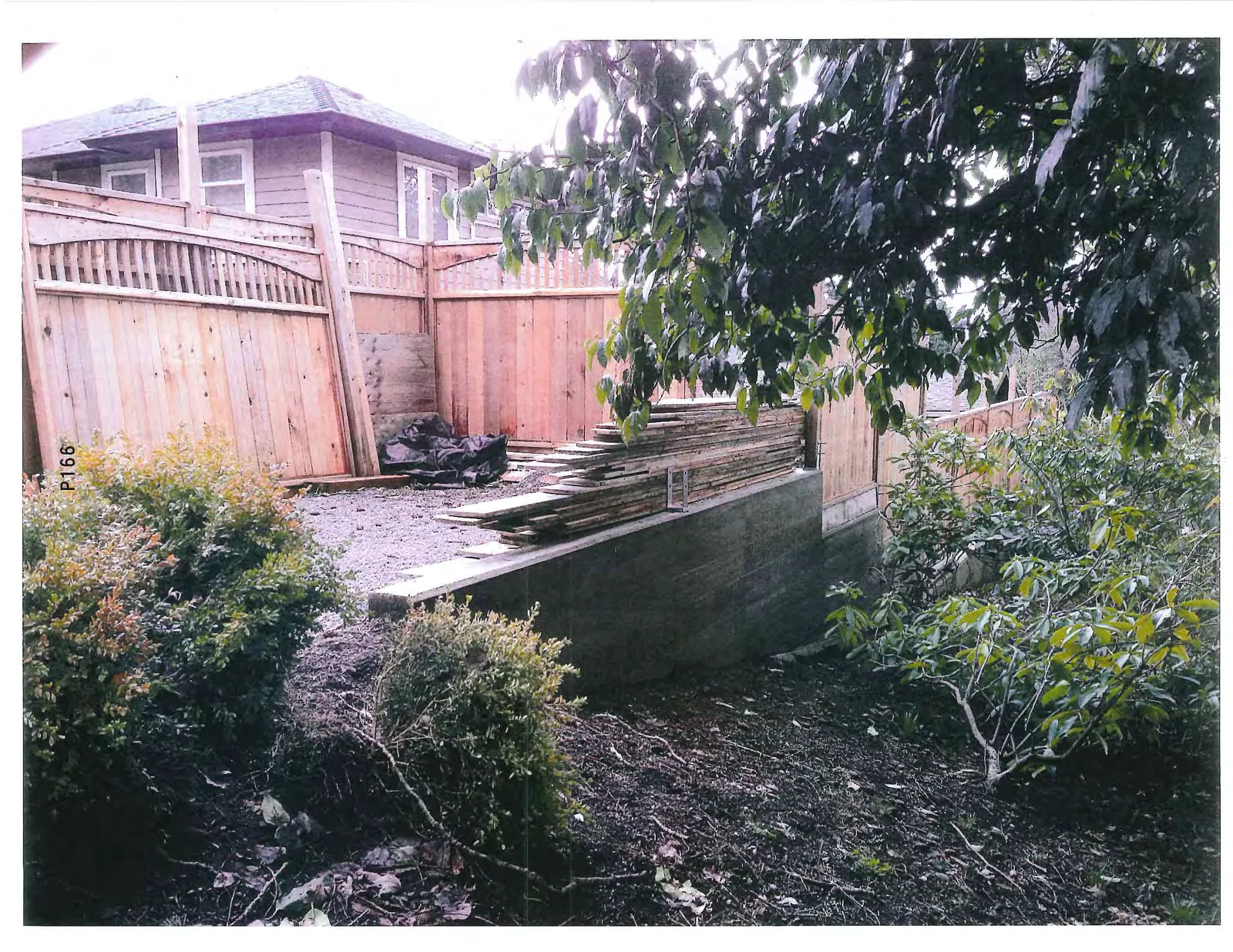Photograph of the wooden fence and concrete retaining wall with stacks of lumber on the ground.