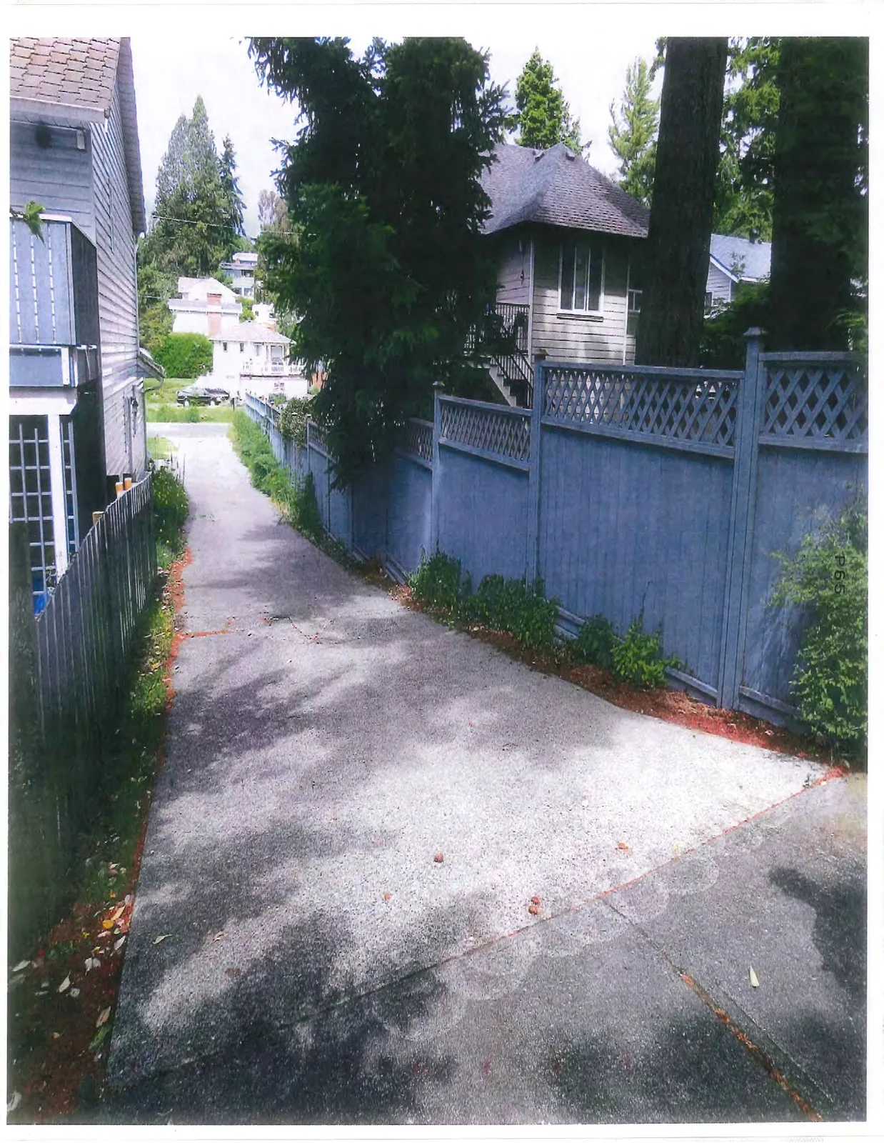 Perspective view of a paved driveway or pathway running alongside a grey wooden fence towards a residential area.
