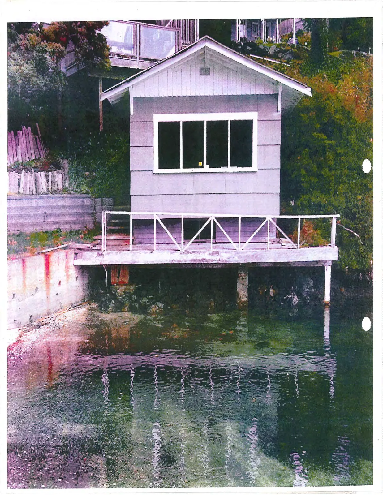 Exterior view of a grey boathouse with white trim and a white wooden deck railing, built over the water.