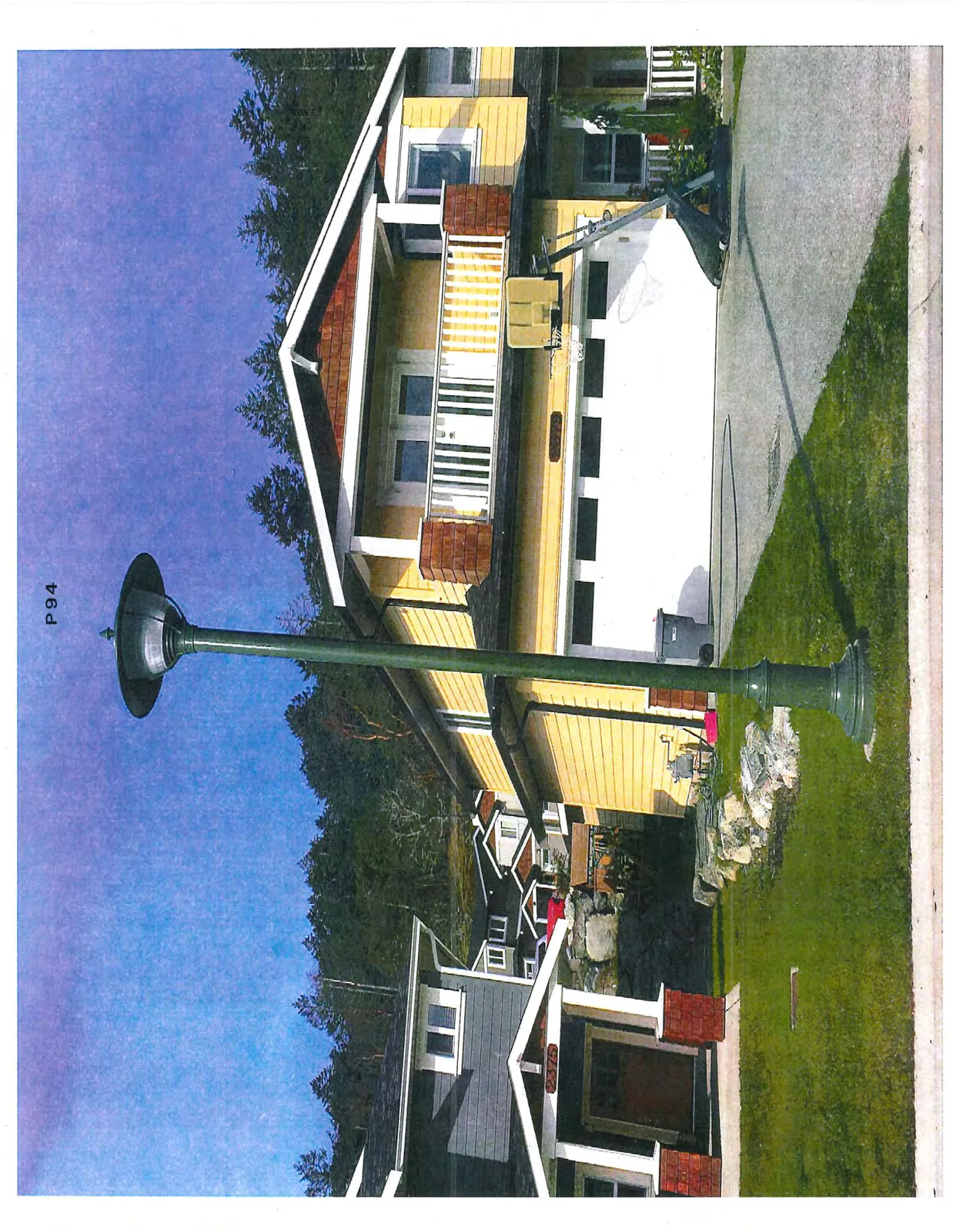 A residential street scene with a prominent green streetlamp in the foreground. On the left is a grey-sided house with the address 2375 above its porch. On the right is a yellow-sided house with a white garage door, address 2379, and a basketball hoop set up in the driveway. The background shows a forested hillside.