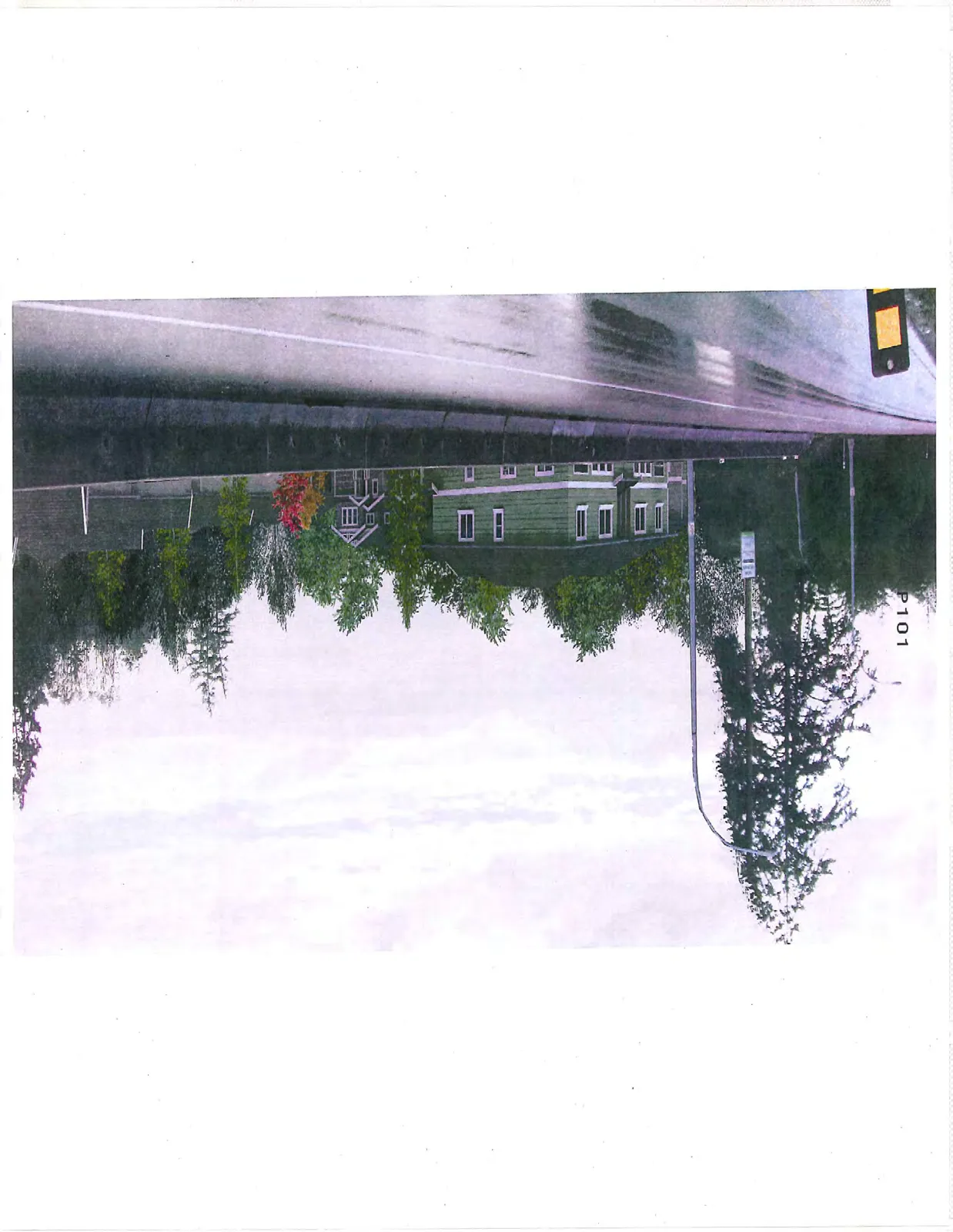 A photograph showing a green multi-story residential building partially obscured by trees, viewed from across a roadway with a concrete barrier.