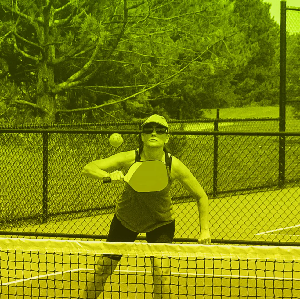 Woman with blonde bun in orange tank top playing pickleball, viewed from behind