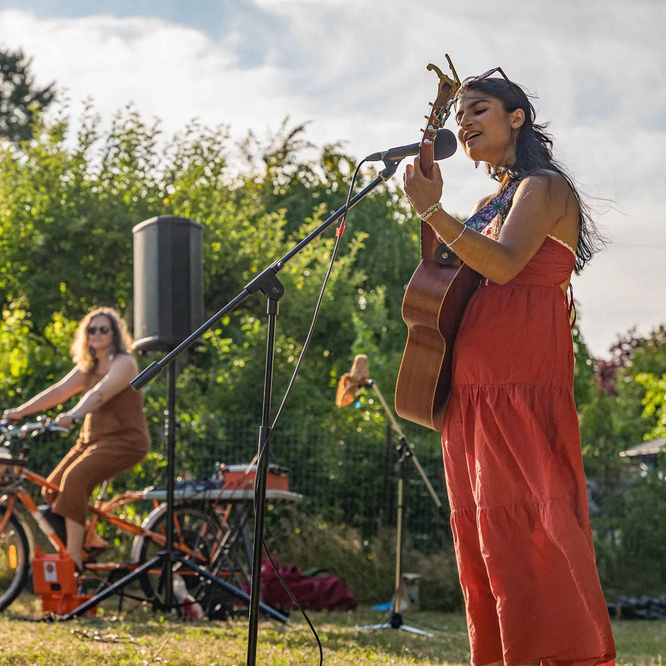 A woman in a red dress playing an acoustic guitar and singing into a microphone outdoors, with another person on a bicycle in the background.