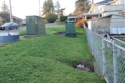 Green utility kiosks on a grass lawn with a red arrow pointing to a drainage manhole