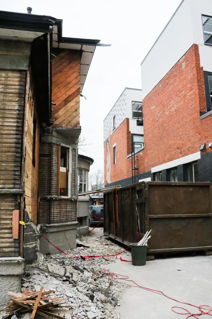 Deconstruction site showing a building partially disassembled next to a newer brick building, with a large waste bin in the foreground