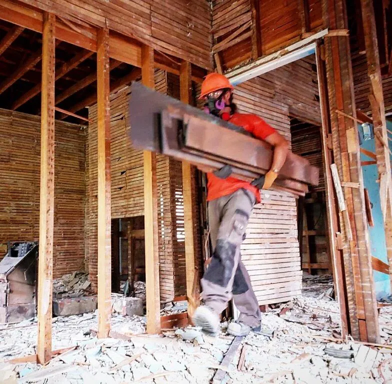 Worker wearing a hard hat and respirator carrying long pieces of salvaged wood through a building under deconstruction