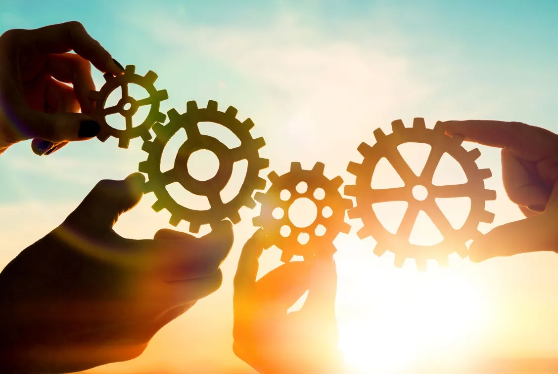 Multiple hands holding interlocking gears together against a bright sky
