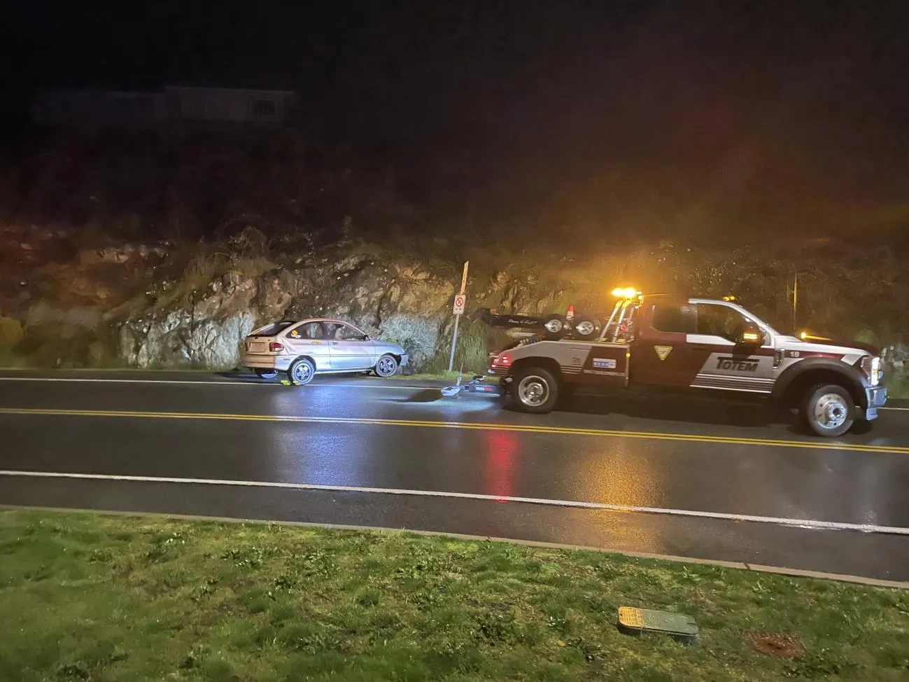 A car accident at night on Watkiss Way where a silver car has crashed into a rock wall next to a wet road, with a Totem tow truck parked nearby.