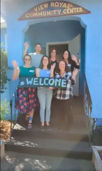 Five members of the View Royal Community Association board waving from the front steps of the center, holding a "WELCOME" sign