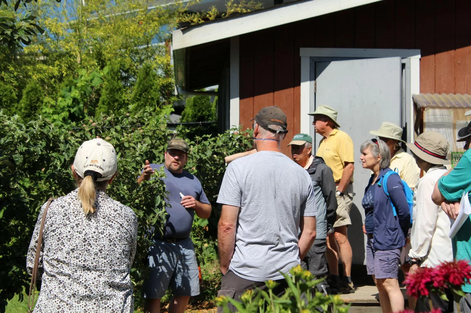 A guide in a blue shirt gesturing toward trees while a group of people in sun hats listens.