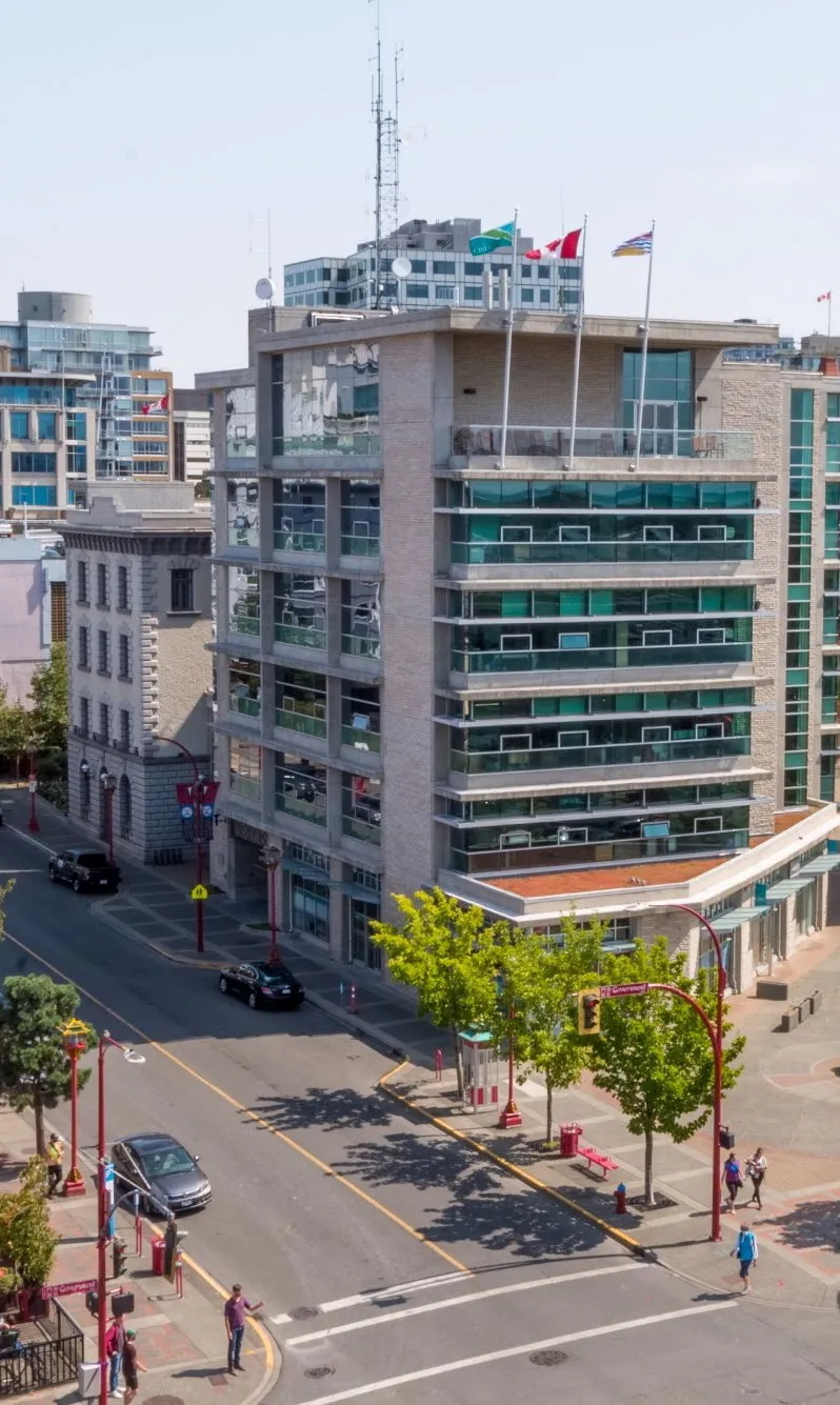 A multi-story modern building with several flags on its roof, situated on a street corner with trees and pedestrians