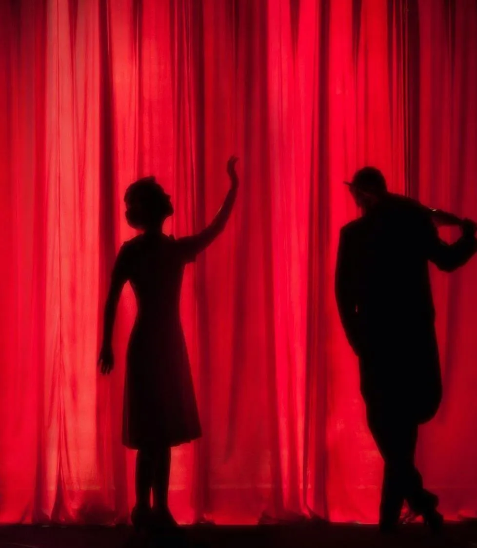 Silhouettes of a man and woman performing on a stage in front of red curtains