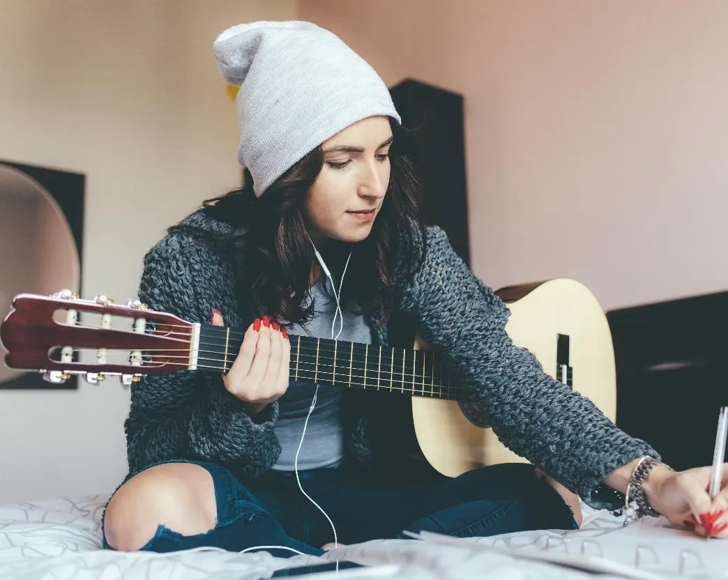 A woman wearing a beanie sitting on a bed playing a guitar and writing on a notepad