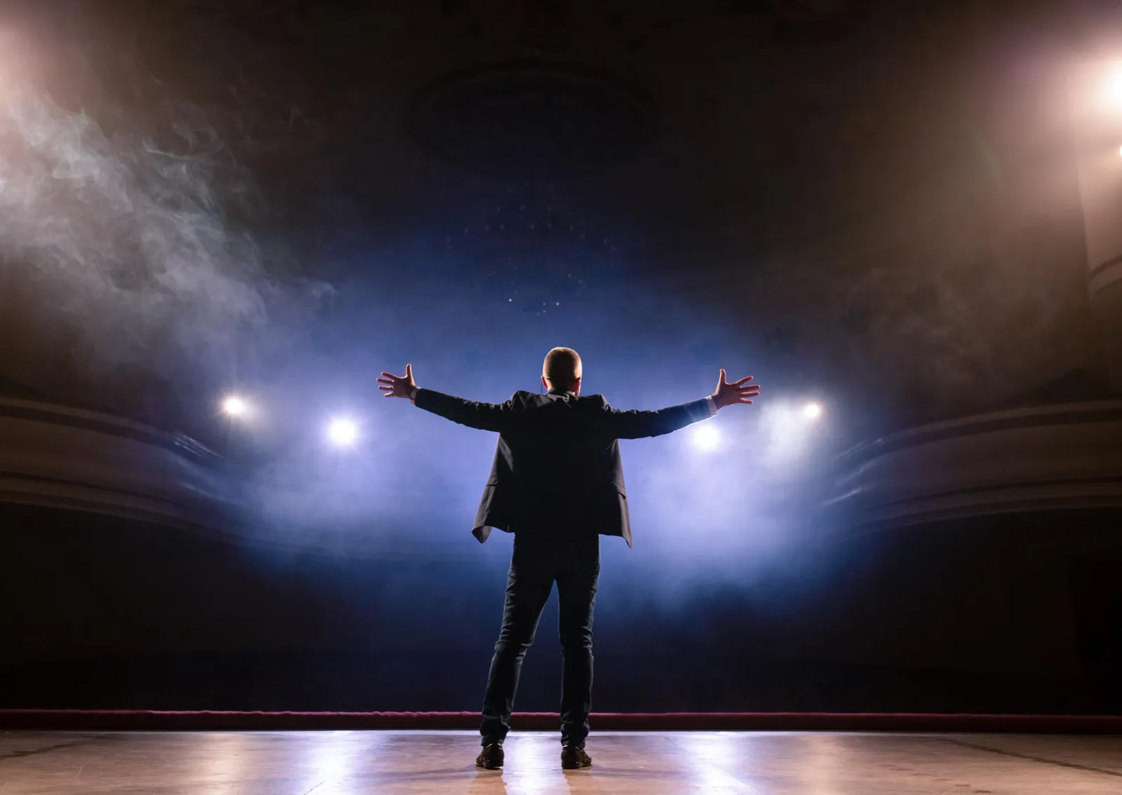 A person standing on a stage with arms outstretched toward bright spotlights