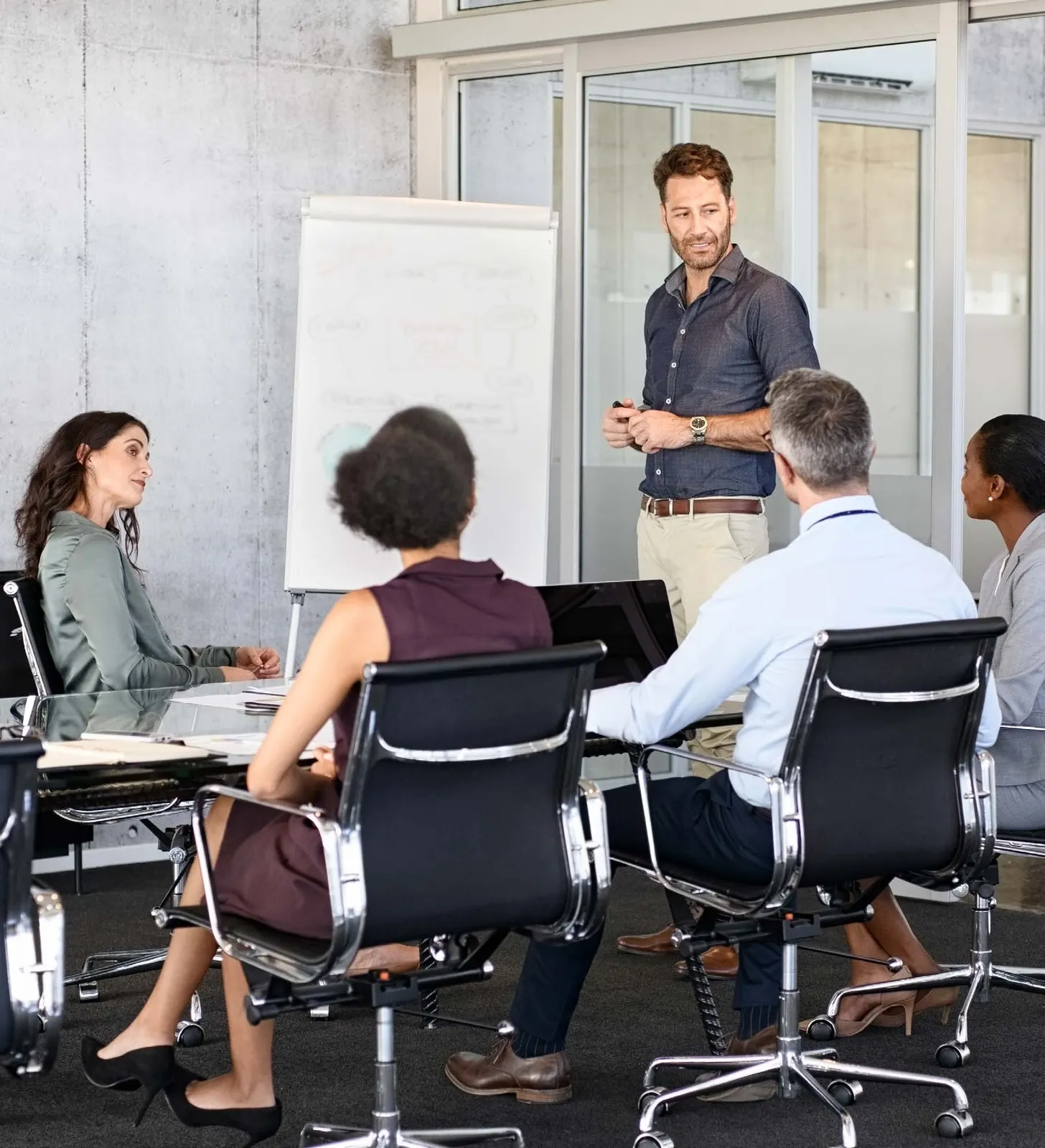 A group of professionals in a meeting room; one man is standing by a whiteboard and presenting to three colleagues seated around a glass conference table.