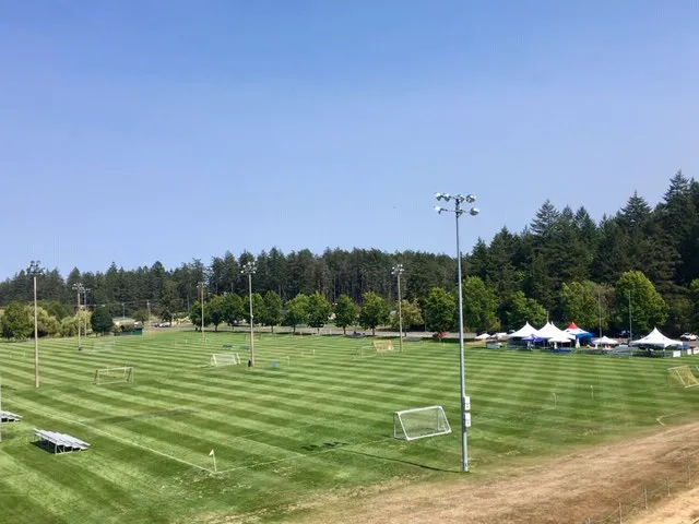Wide-angle photograph of well-maintained outdoor athletic fields with goalposts, light towers, and surrounding trees under a clear blue sky