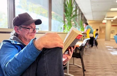 Man in a hat reading a book in a sunlit library aisle