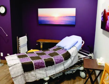 A clinic room with a medical bed, a purple wall, and a sunset landscape painting