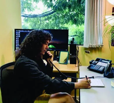 A staff member with curly hair sitting at a desk, talking on a telephone and taking notes