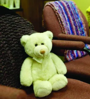 A yellow teddy bear on a brown chair next to a colourful knitted blanket