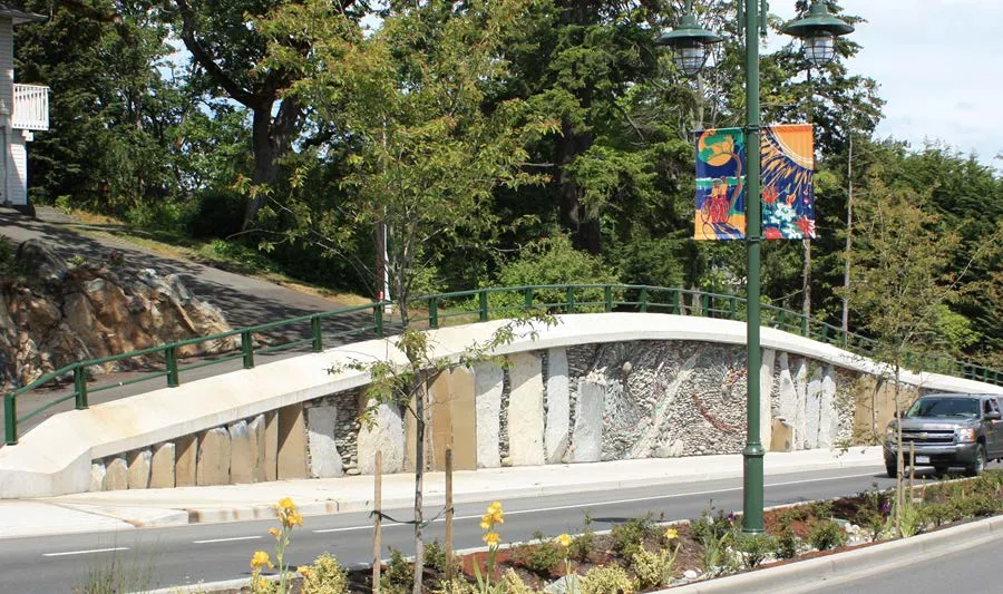 Photograph of a bridge wall featuring vertical stone slabs and mosaic-style artwork, with colorful art banners hanging from a lamppost in front of it.