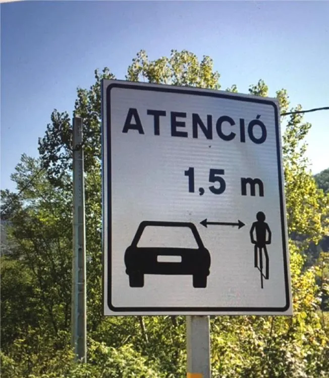 A roadside sign that reads "ATENCIÓ 1,5 m" with a graphic of a car and a cyclist separated by a double-headed arrow.