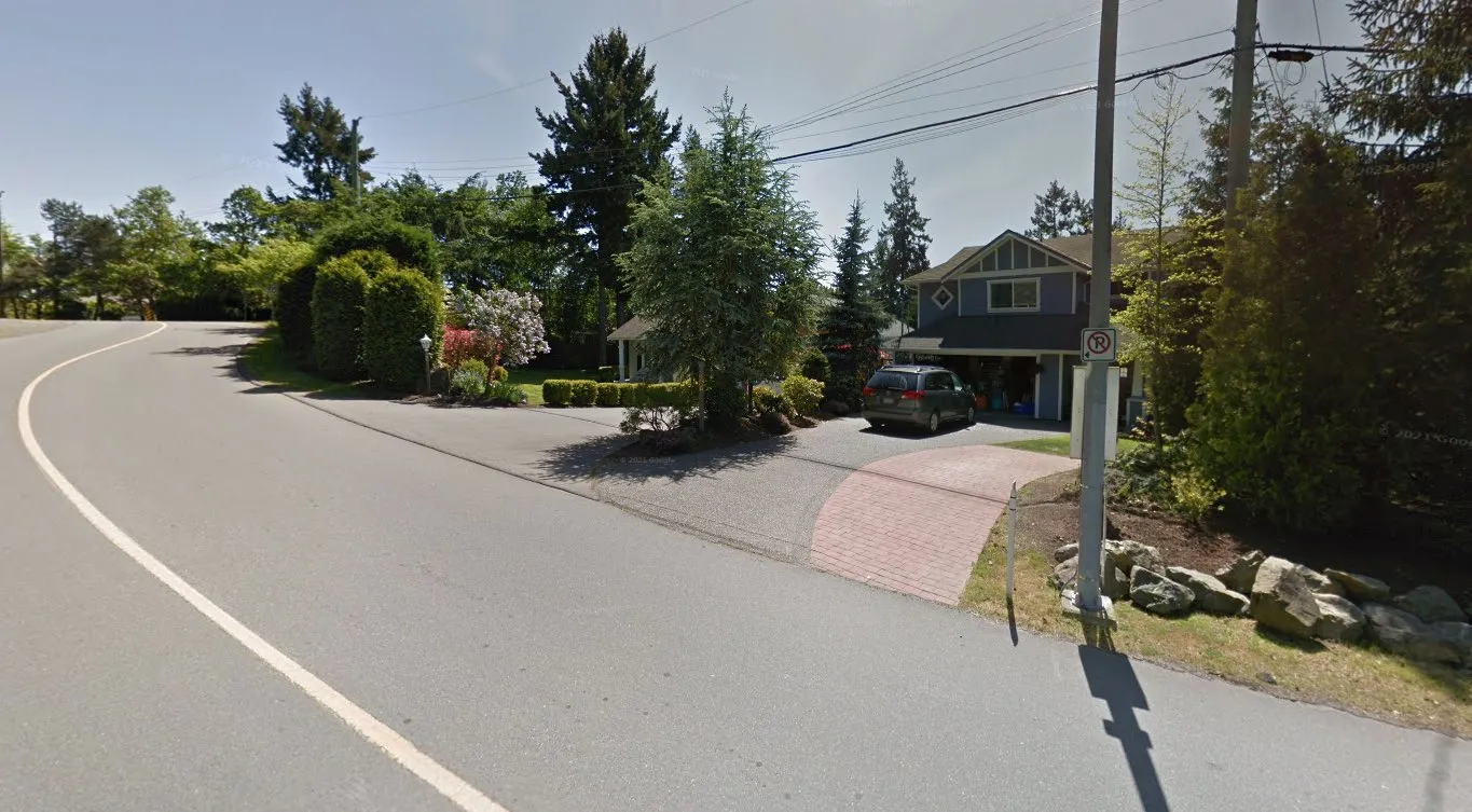 Street view photo showing a blue two-story house with a grey minivan parked in the driveway, situated on a curved road with lush green trees and landscaping.