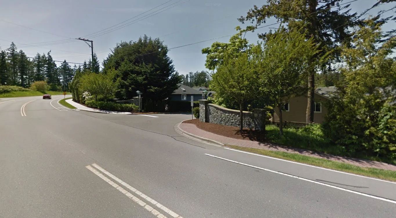 A street-level photograph showing a paved road curving to the left with a red car in the distance. In the foreground, a stone wall marks the entrance to a residential area, surrounded by trees and green foliage.