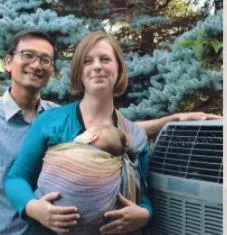 A man and a woman holding a baby in a wrap standing next to an outdoor heat pump unit.