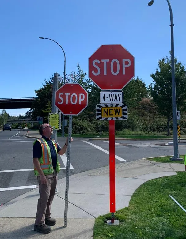 A worker in a high-visibility vest standing next to a permanent stop sign that includes "4-WAY" and "NEW" placards, while holding up a second stop sign on a tall post.