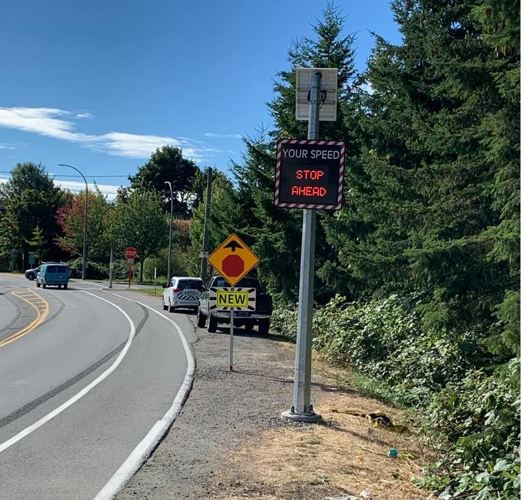 A roadside view showing the "YOUR SPEED / STOP AHEAD" digital sign and a yellow warning sign with a stop sign symbol and a "NEW" placard.