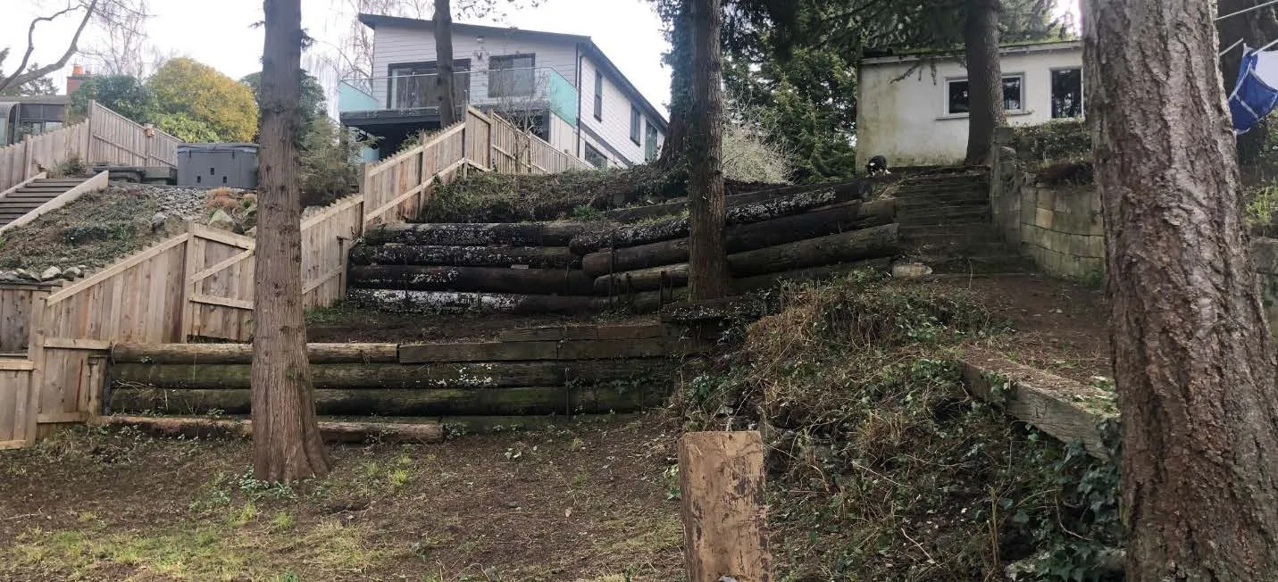 Photograph of the tiered backyard area featuring old wooden log retaining walls and stairs