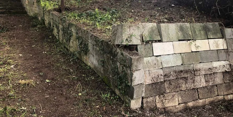 Photograph showing a damaged or aged concrete block retaining wall with vegetation growing through cracks
