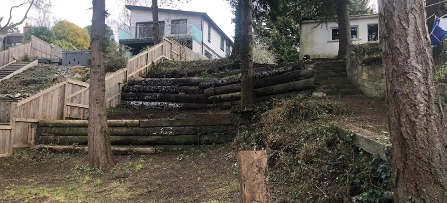 Photograph showing tiered wooden retaining walls made of logs on a sloped, dirt-covered property with houses in the background