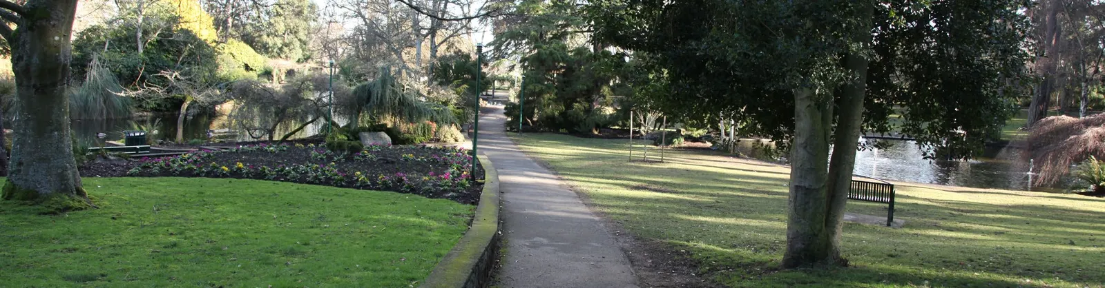 A panoramic photo of a park with a paved path, lawn, trees, and a pond.