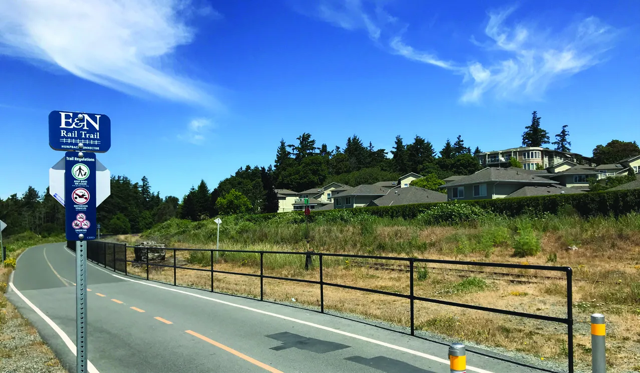 A view of the E&N Rail Trail path with a sign for trail regulations and houses in the background under a blue sky.