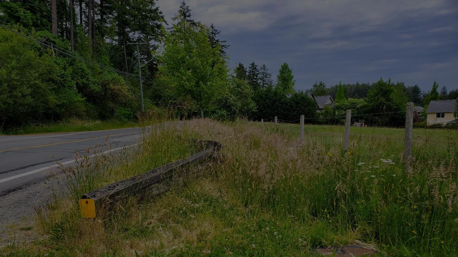 View of Atkins Road showing a grassy shoulder, a split-rail wooden fence, and tall grass in the foreground