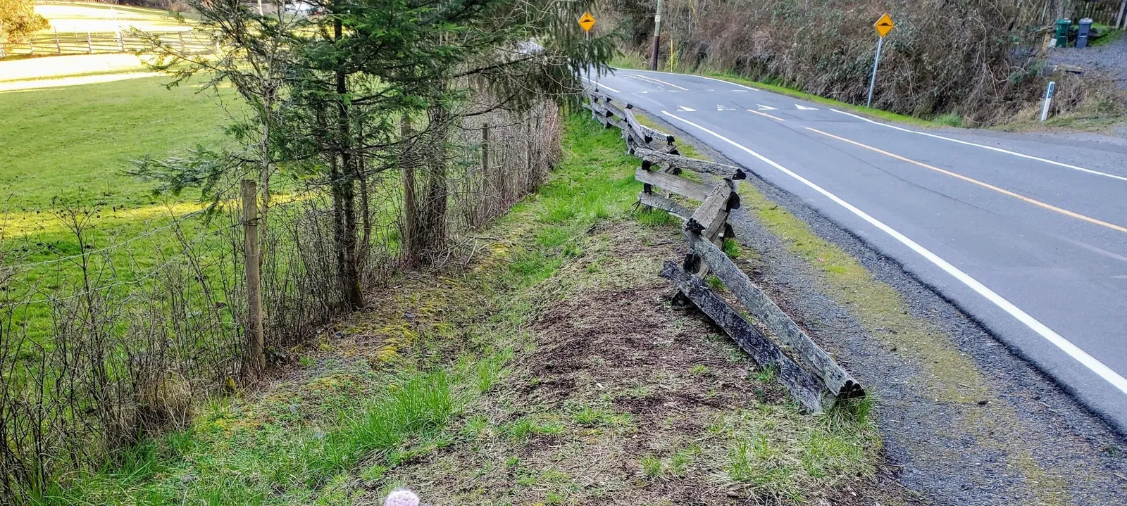 Narrow road shoulder on Atkins Road situated between the paved traffic lane and a weathered wooden fence