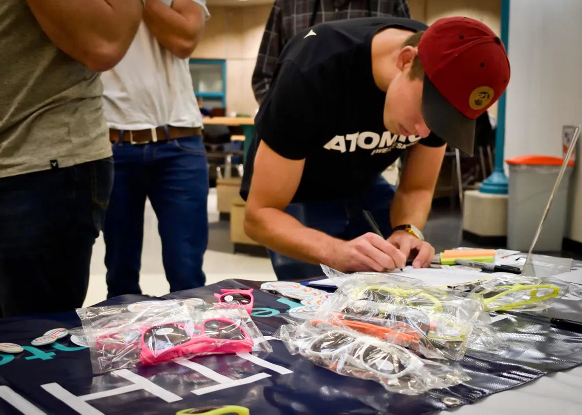 Man in a black "ATOMIC" shirt writing at a table with colorful sunglasses in bags