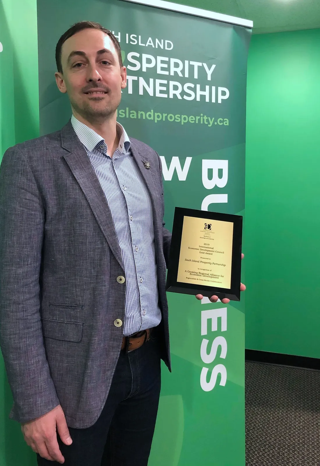 Man in a blazer holding an award plaque in front of a "South Island Prosperity Partnership" banner