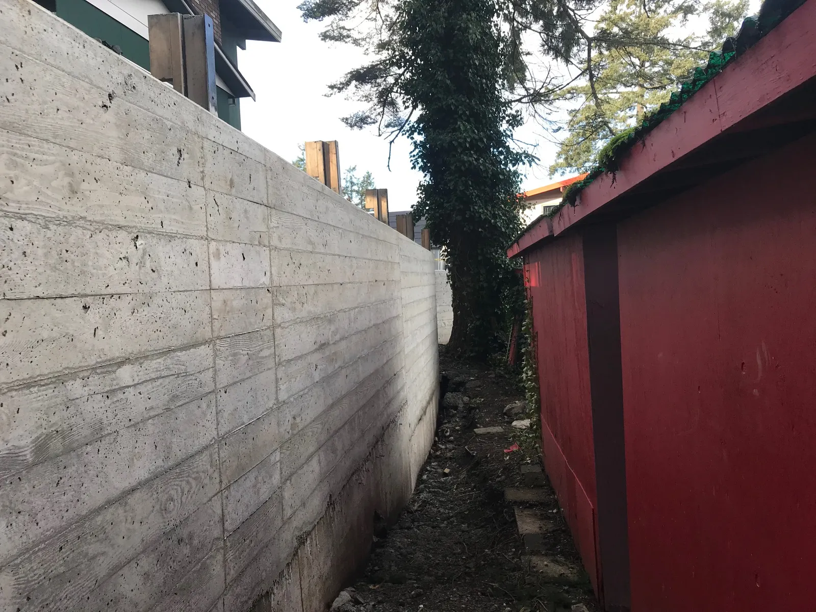 Narrow pathway between the new concrete wall and a red wooden structure.