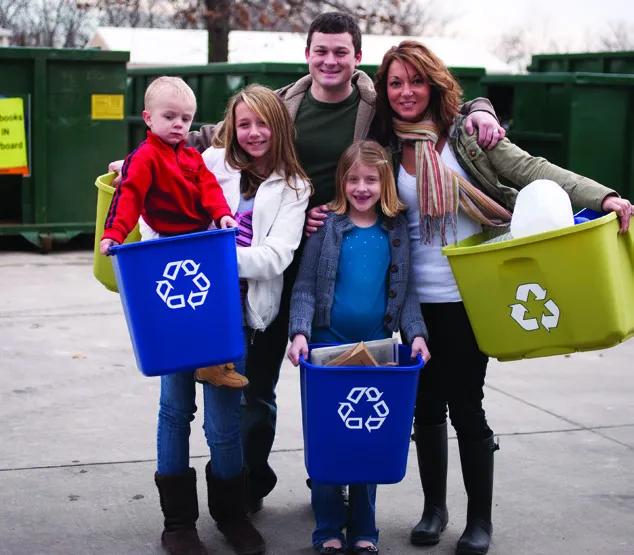 A family standing in front of large recycling bins holding their own blue bins.
