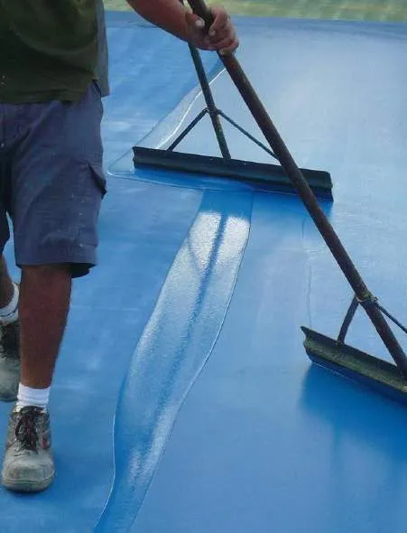 A worker using a large squeegee to apply a blue coating to a court surface