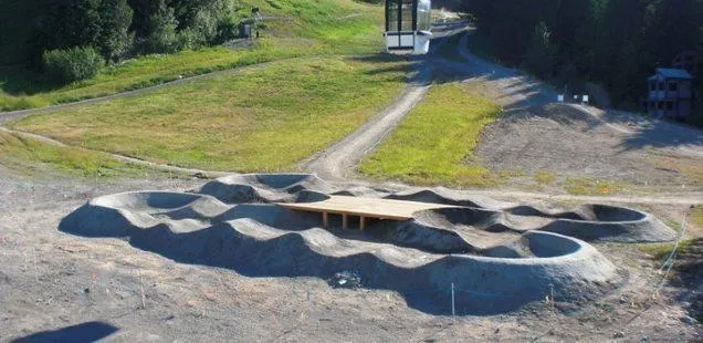 aerial view of a paved pump track on a slope near a ski lift, featuring a wooden center platform