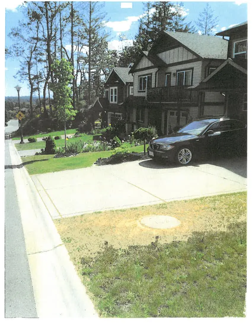 A photograph of a residential street scene. In the foreground, there is a patch of lawn with dry, yellowed grass containing a circular manhole cover. A concrete driveway is to the right of the lawn, where a black car is parked in front of a row of two-story houses with tan siding and dark trim. To the left, a sidewalk runs downhill alongside a paved road, passing a street light with a yellow pedestrian crossing sign.