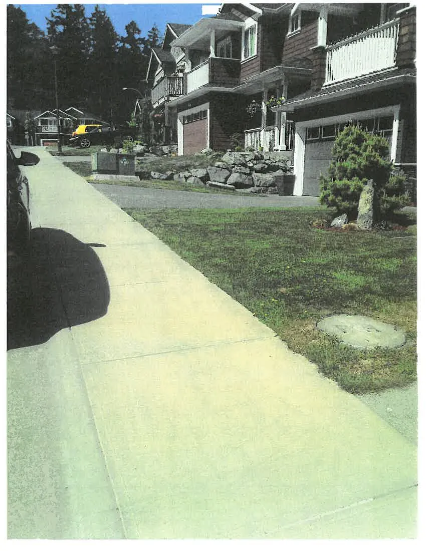 A photograph of a residential area viewed from a sidewalk. The concrete sidewalk leads into the distance, with a grassy area on the right that features a circular manhole cover and a small evergreen tree. In the background, several houses with garage doors and a tiered rock retaining wall are visible. A black car is partially visible on the left side of the frame, and a yellow vehicle is parked further down the street.