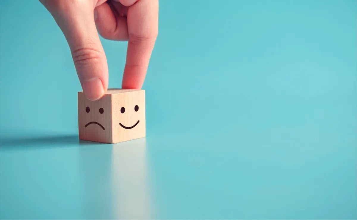 Two fingers resting on a wooden block that shows a frowny face on one side and a smiley face on the other