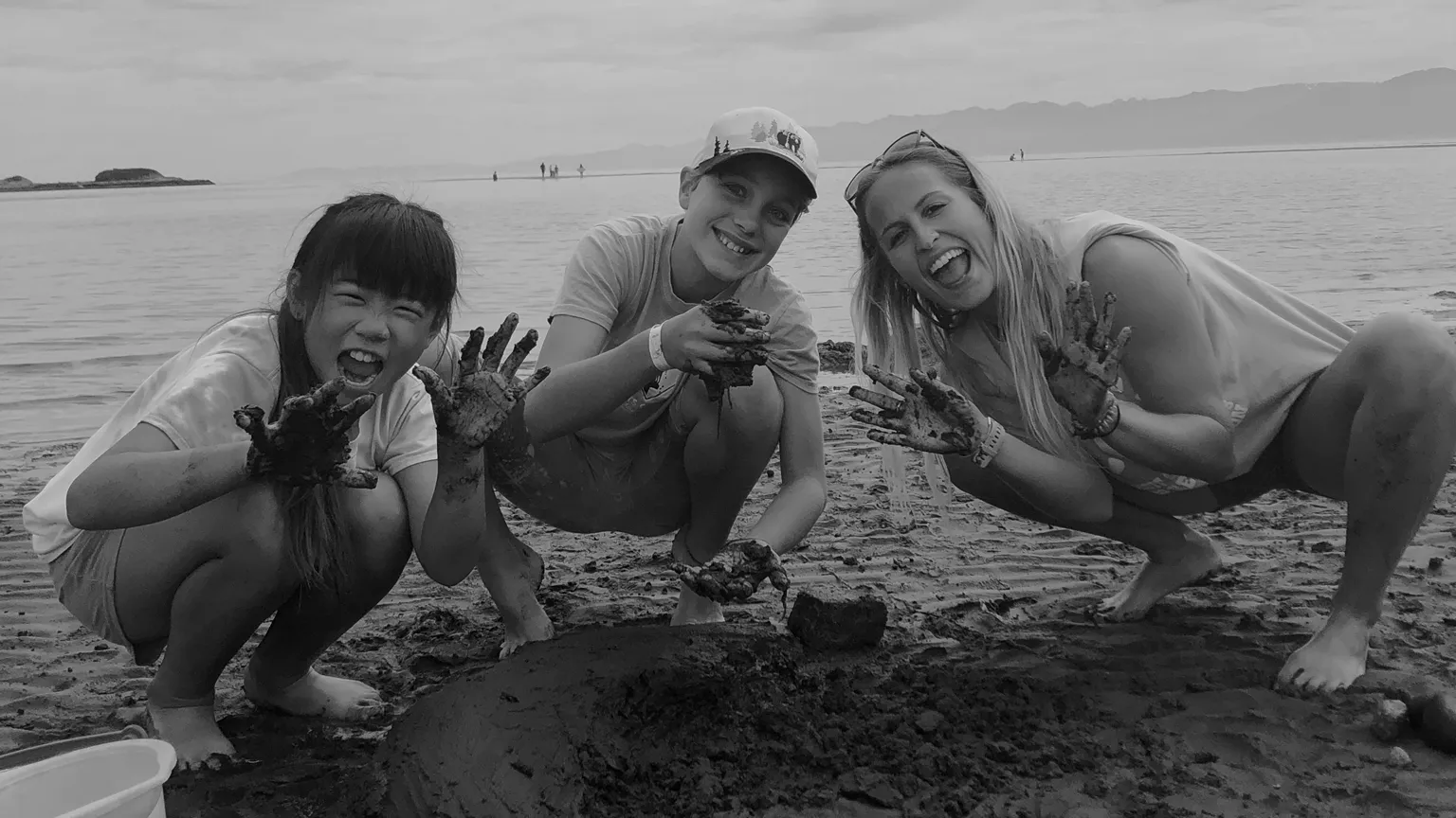 Three people smiling and showing their mud-covered hands at a beach