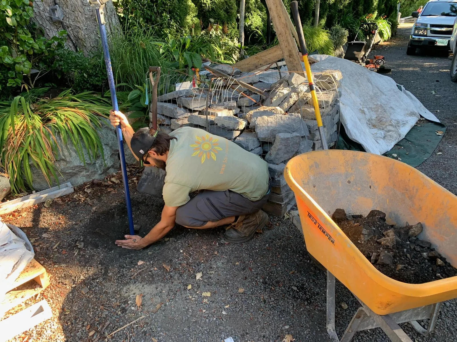 A man in a green shirt kneeling on a gravel driveway, using a long blue-handled tool to probe a small hole, next to an orange wheelbarrow and a wire-caged rock wall.