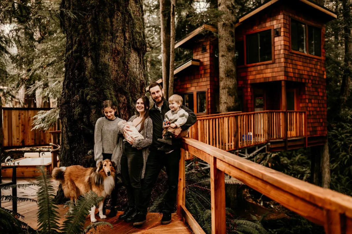 A family with a baby and a dog standing on a wooden walkway in front of a shingled treehouse-style home in a forest setting